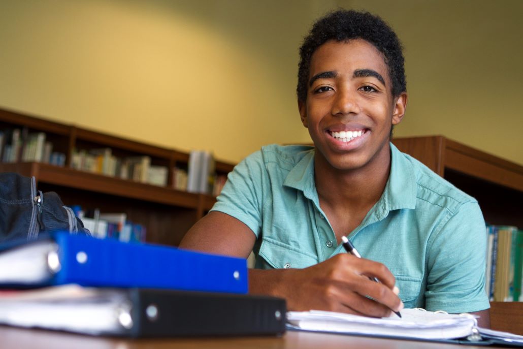 Teenage boy taking notes while applying to college in the library