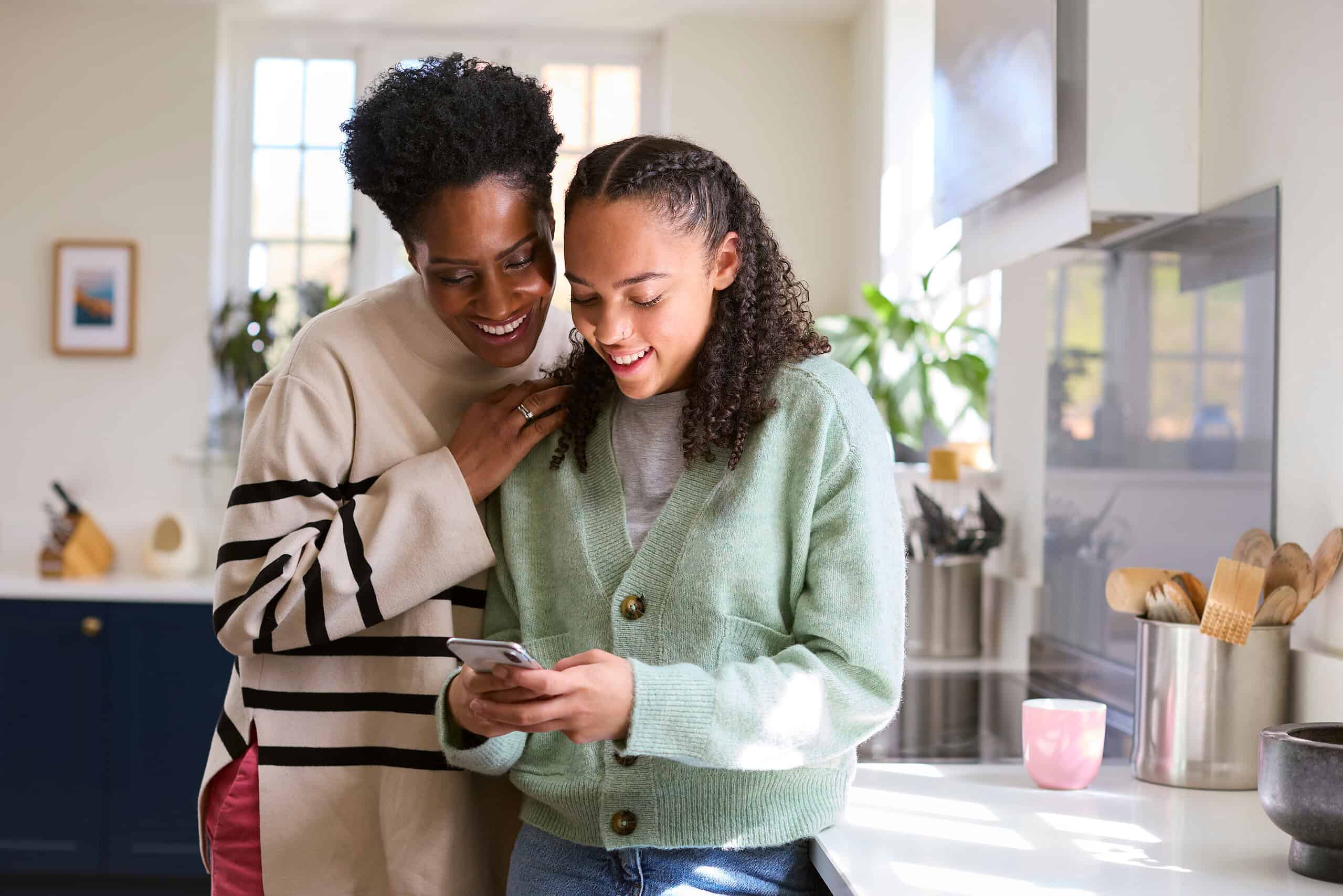 Mother Talking With Teenage Daughter At Home Looking At Social Media On Mobile Phone Together